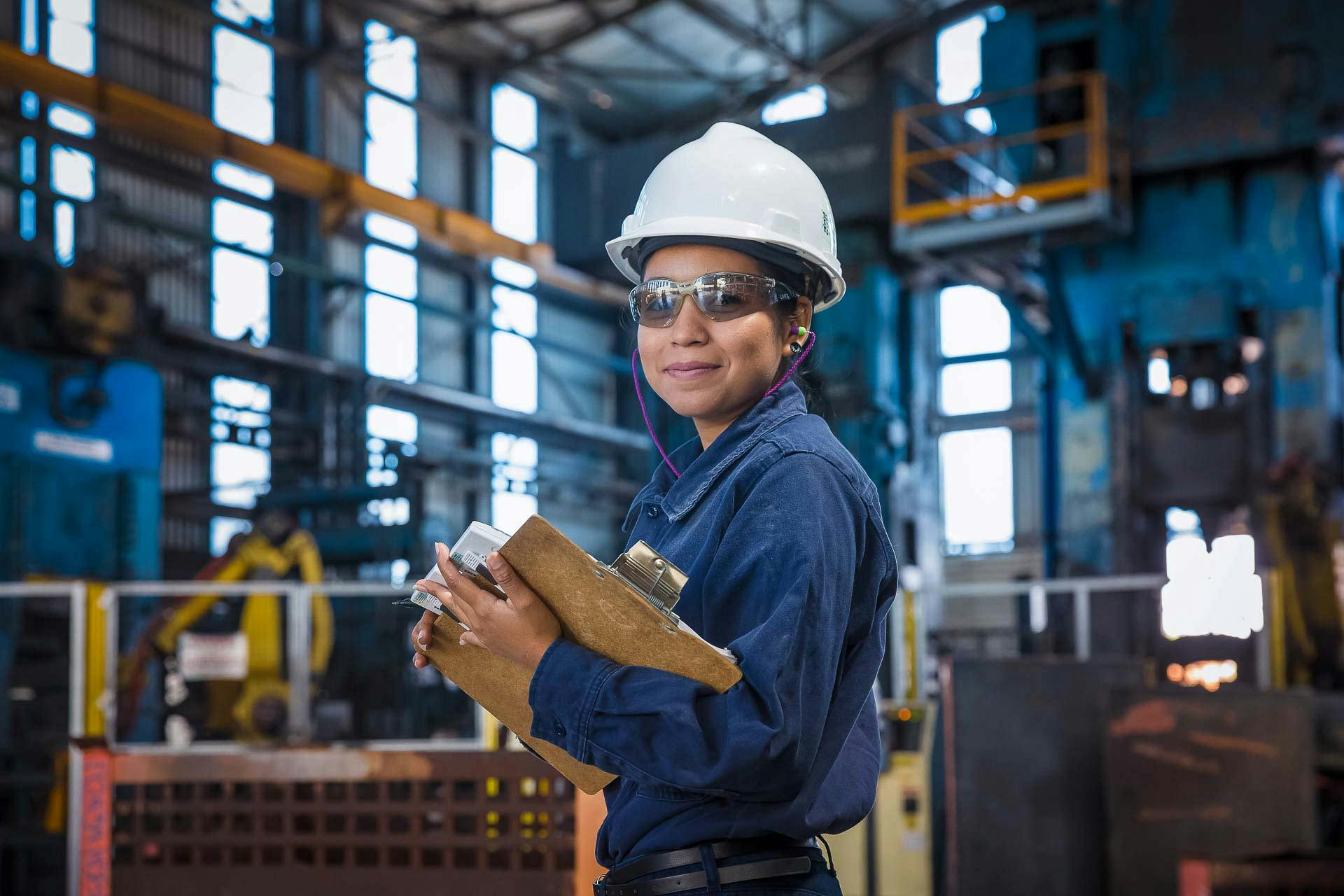 A worker smiles while holding a clipboard in a manufacturing facility