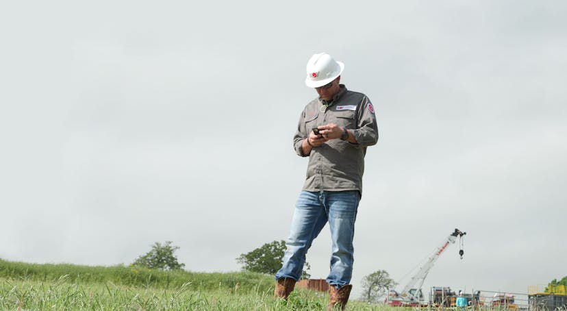 Max app user on his phone while standing in a field with a NOV hard hat on