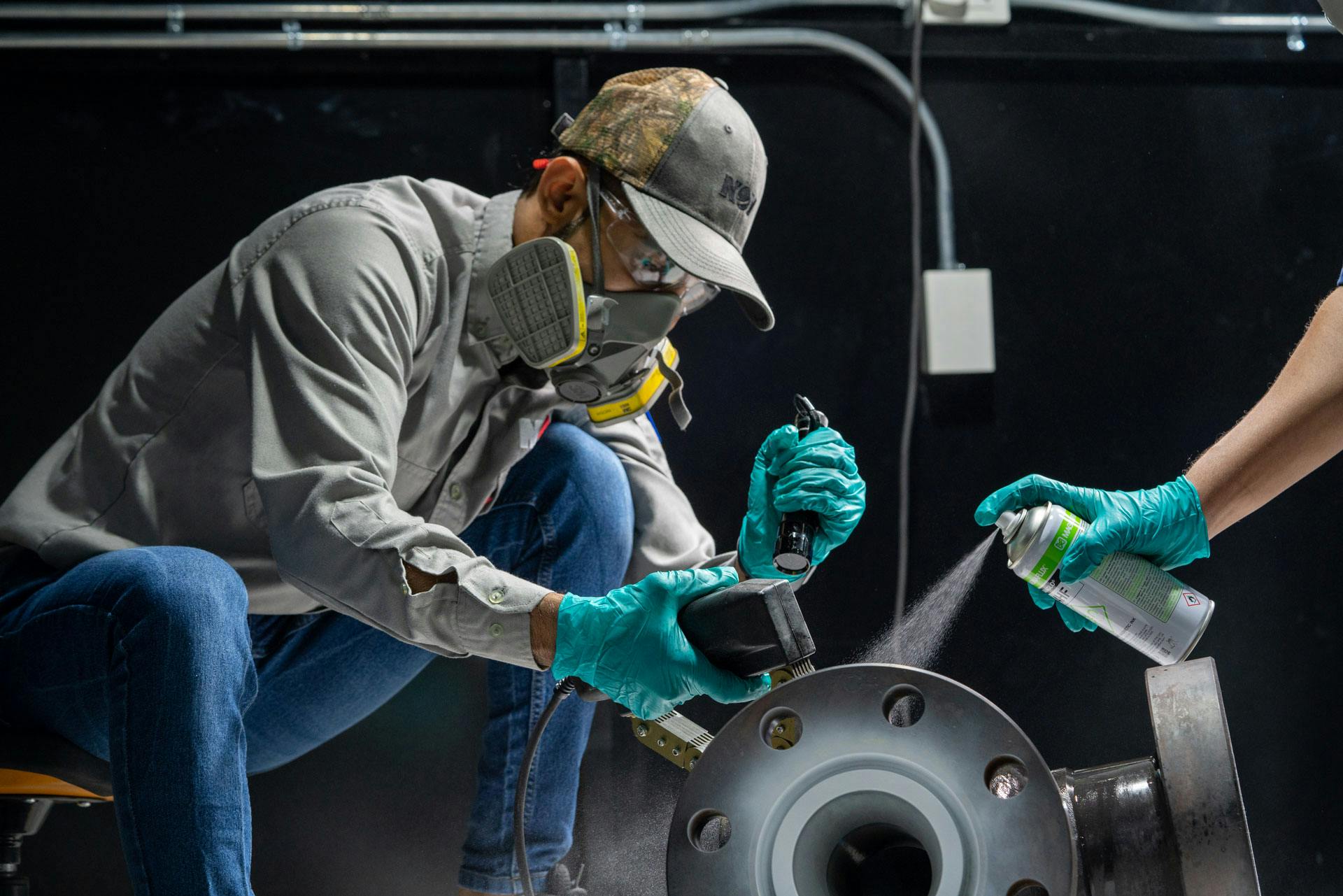 NOV employee wearing PPE while painting equipment, with a hand entering the shot with a spray can