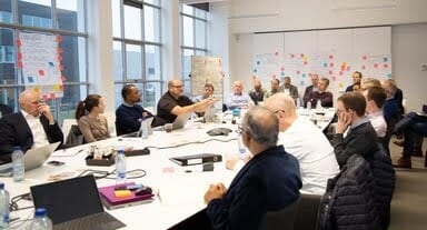 Group of people meeting and discussing around a large table in a modern office with laptops.