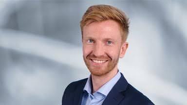 Smiling man with red hair and beard wearing a blue shirt and dark suit jacket.