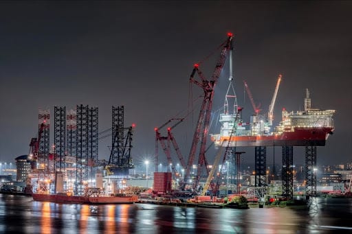 Night view of an industrial port with large ships, cranes, and lights reflecting on water.