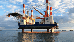 Offshore jack-up rig with red and white striped legs deployed, hull lifted above water.