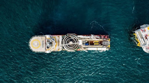 Aerial view of a pipe-laying vessel with helipad, next to another ship on blue water.