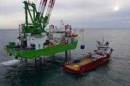 Green jack-up vessel lifting cargo from red supply ship offshore.