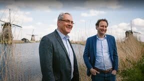 Two smiling men in a Dutch landscape with windmills and a canal.