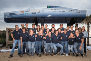 Group in blue shirts poses with a high-standing boat on a beach.