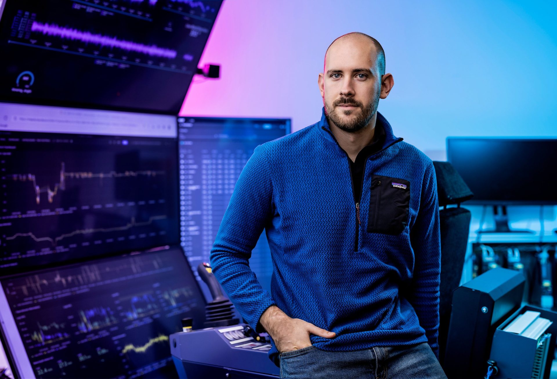 Man in a blue sweater by a multi-monitor setup displaying data with purple and blue lighting.
