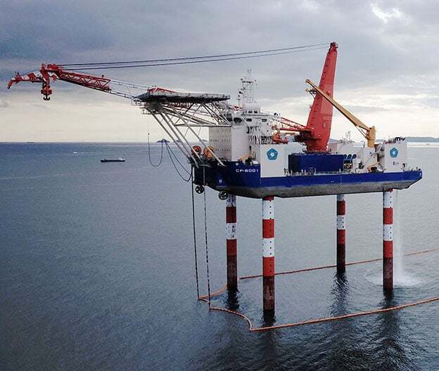 Jack-up vessel on striped legs with two cranes in the ocean under a cloudy sky.