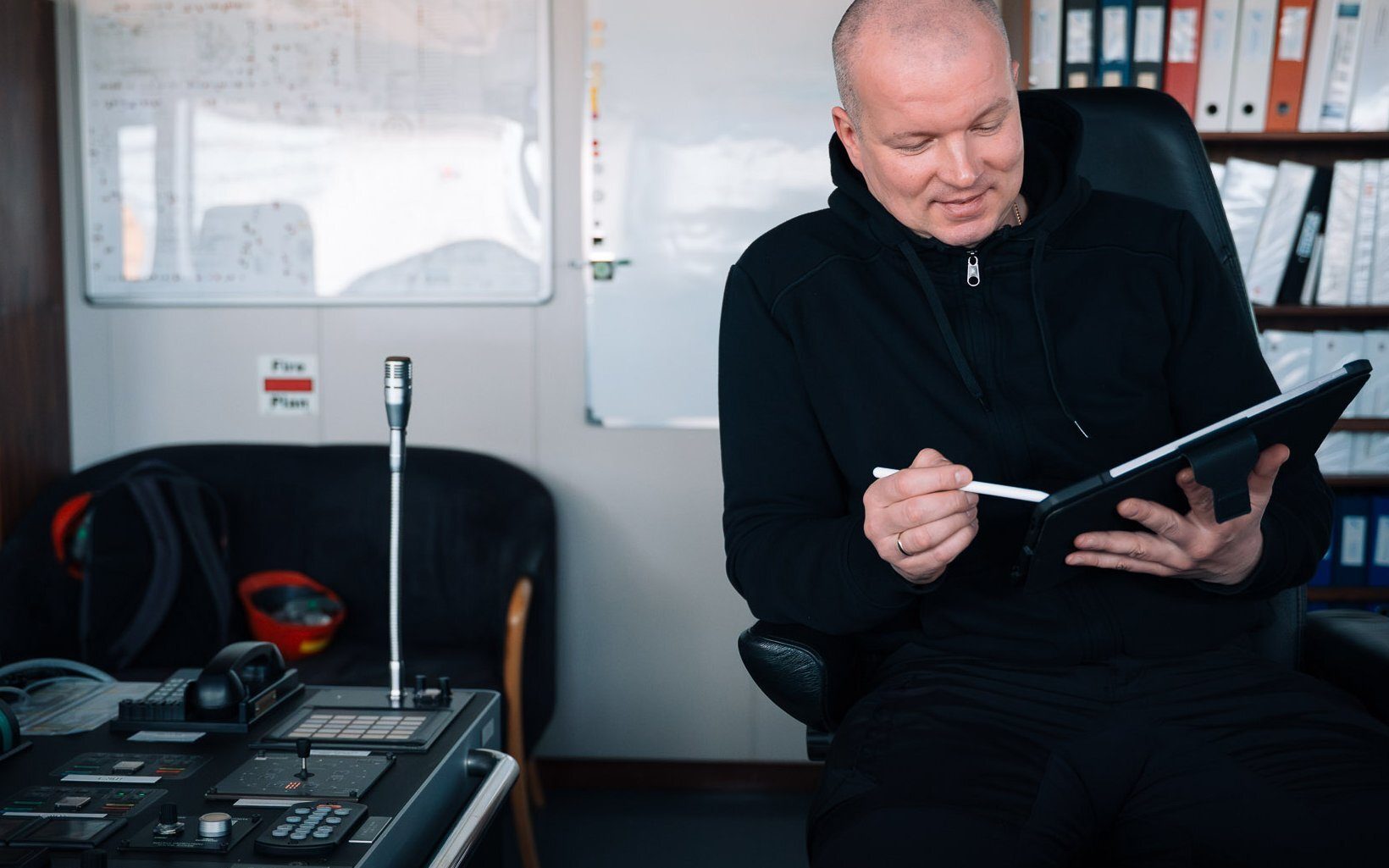 Man in black hoodie using a tablet with stylus in a control room.