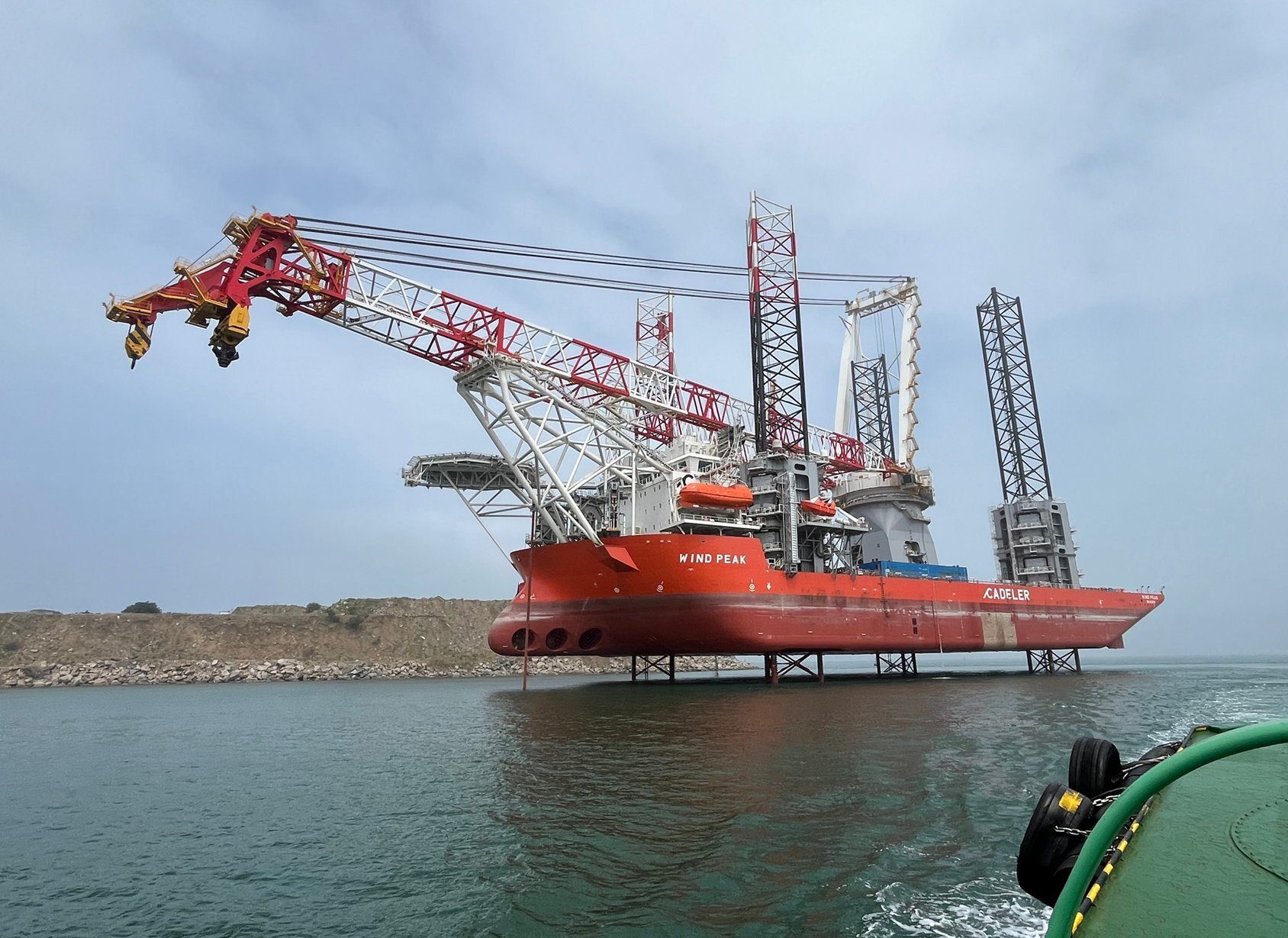 Red jack-up vessel 'WIND PEAK' with cranes, elevated above water near shore.