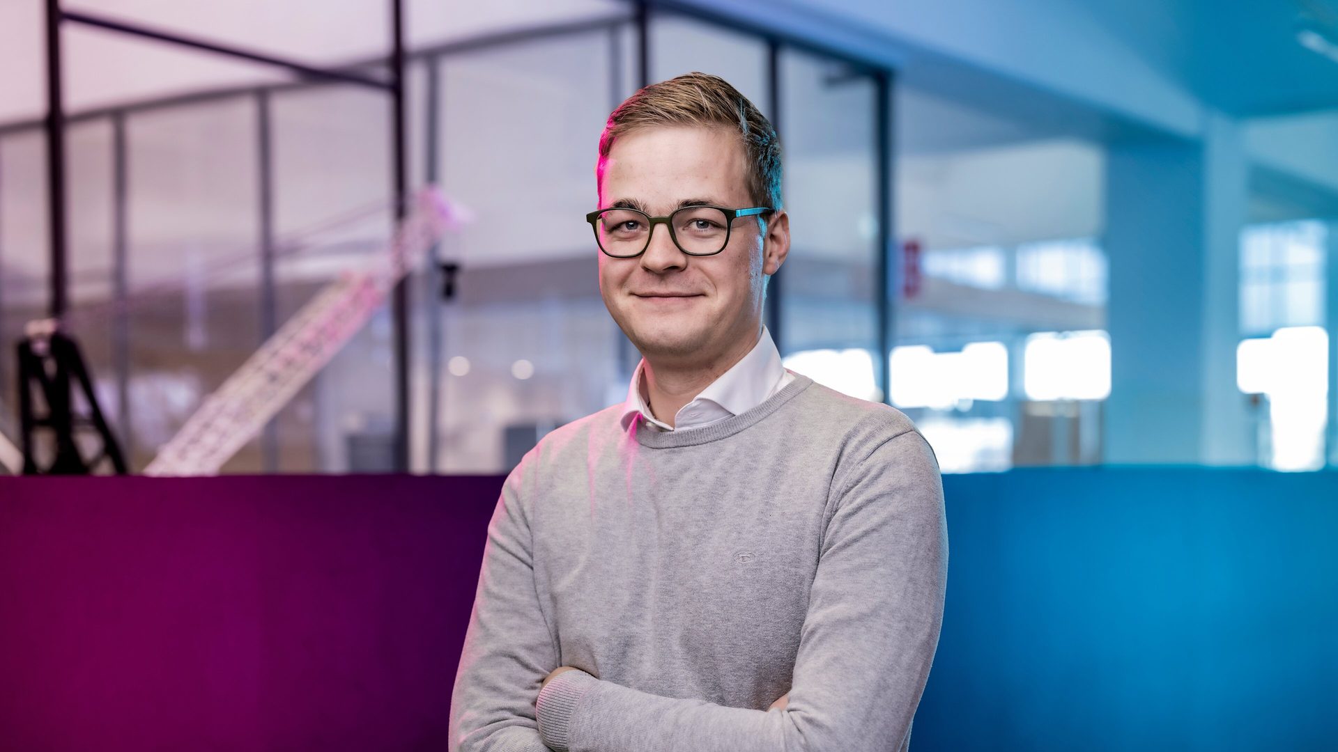Smiling man in glasses and gray sweater, arms crossed. Blurred modern office background.