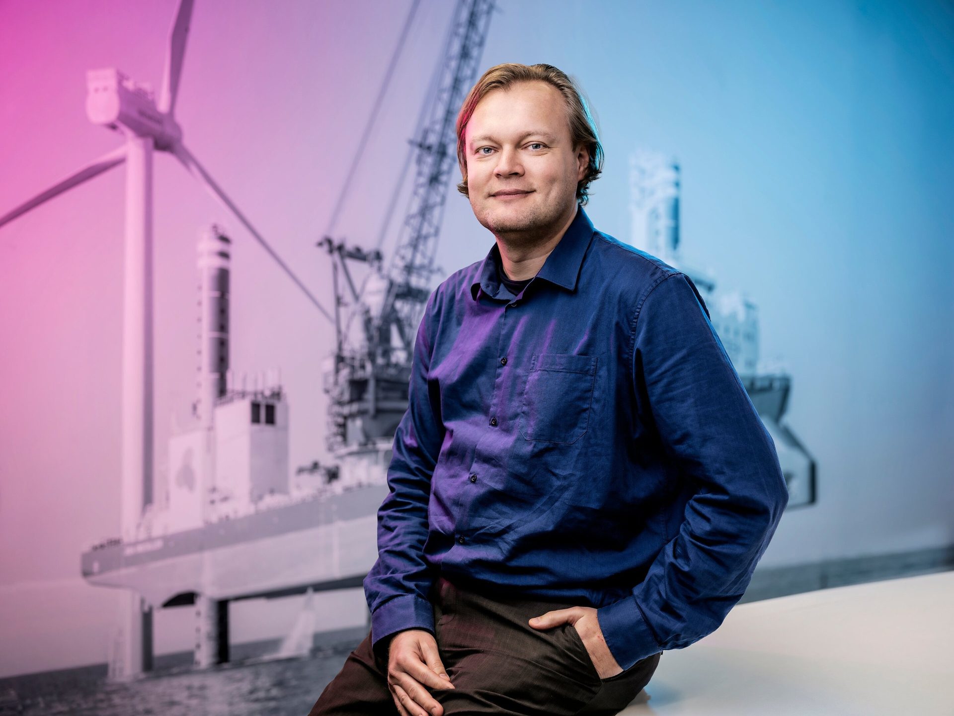 Smiling man in blue shirt before a blurred, pink-blue industrial background.