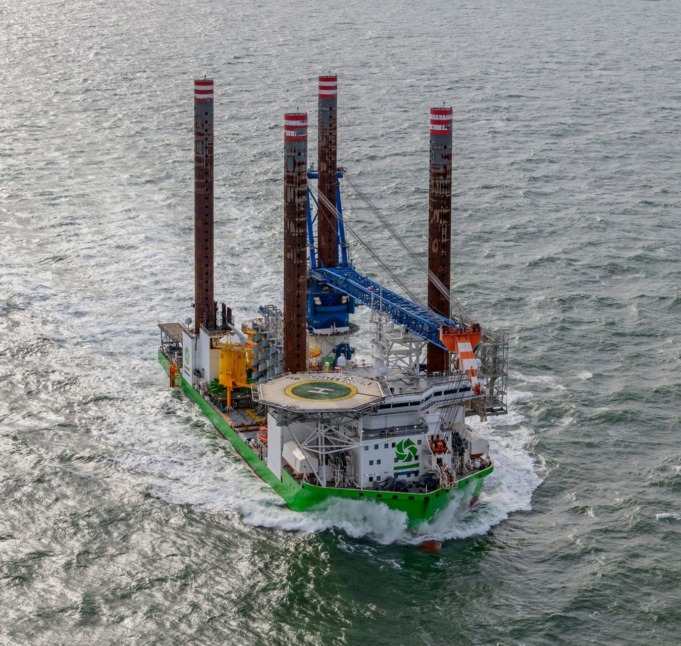 Green jack-up vessel with four tall legs and a crane, leaving a wake in choppy water.