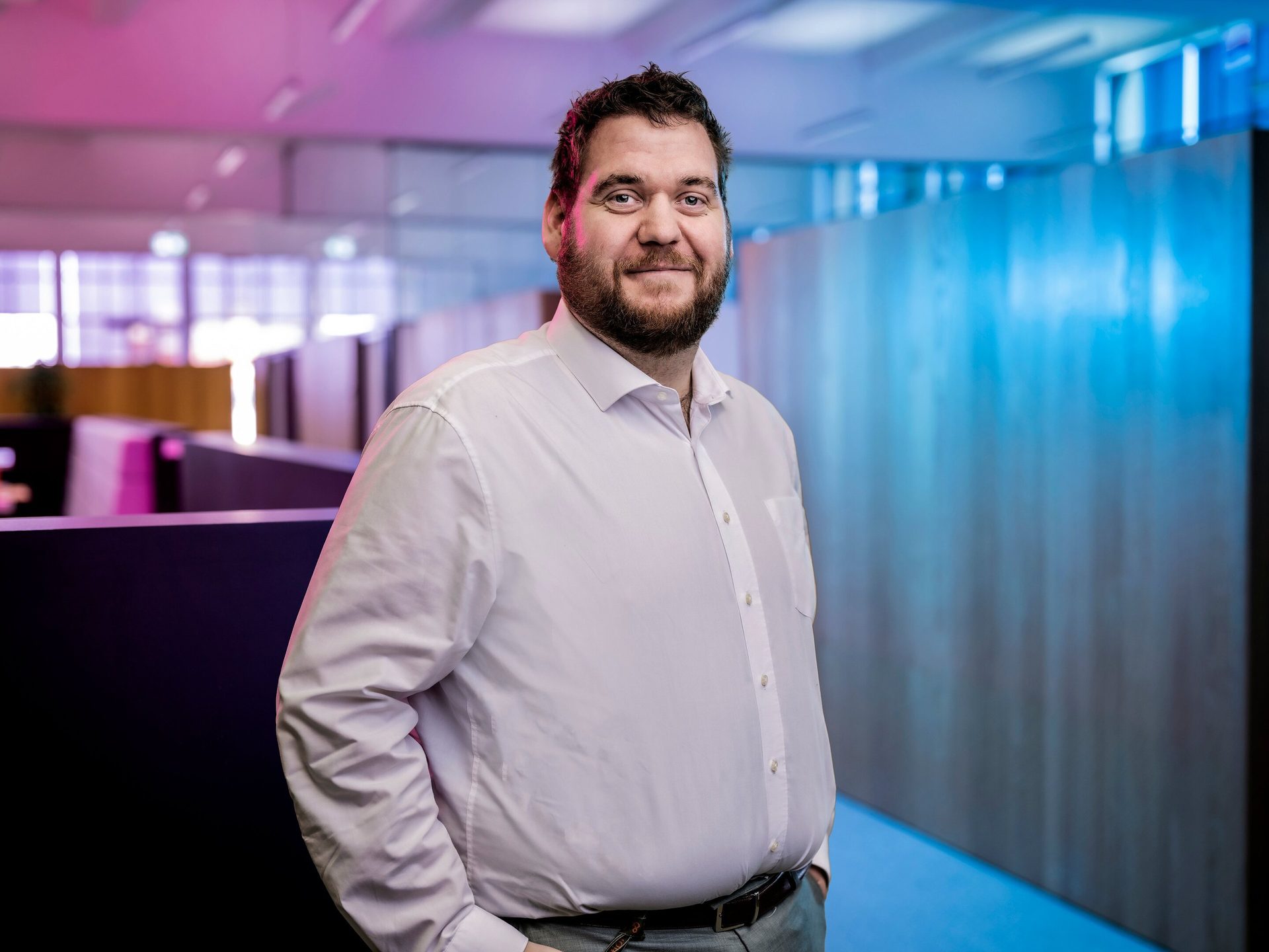 A smiling bearded man in a white shirt, standing in an office with pink and blue lighting.
