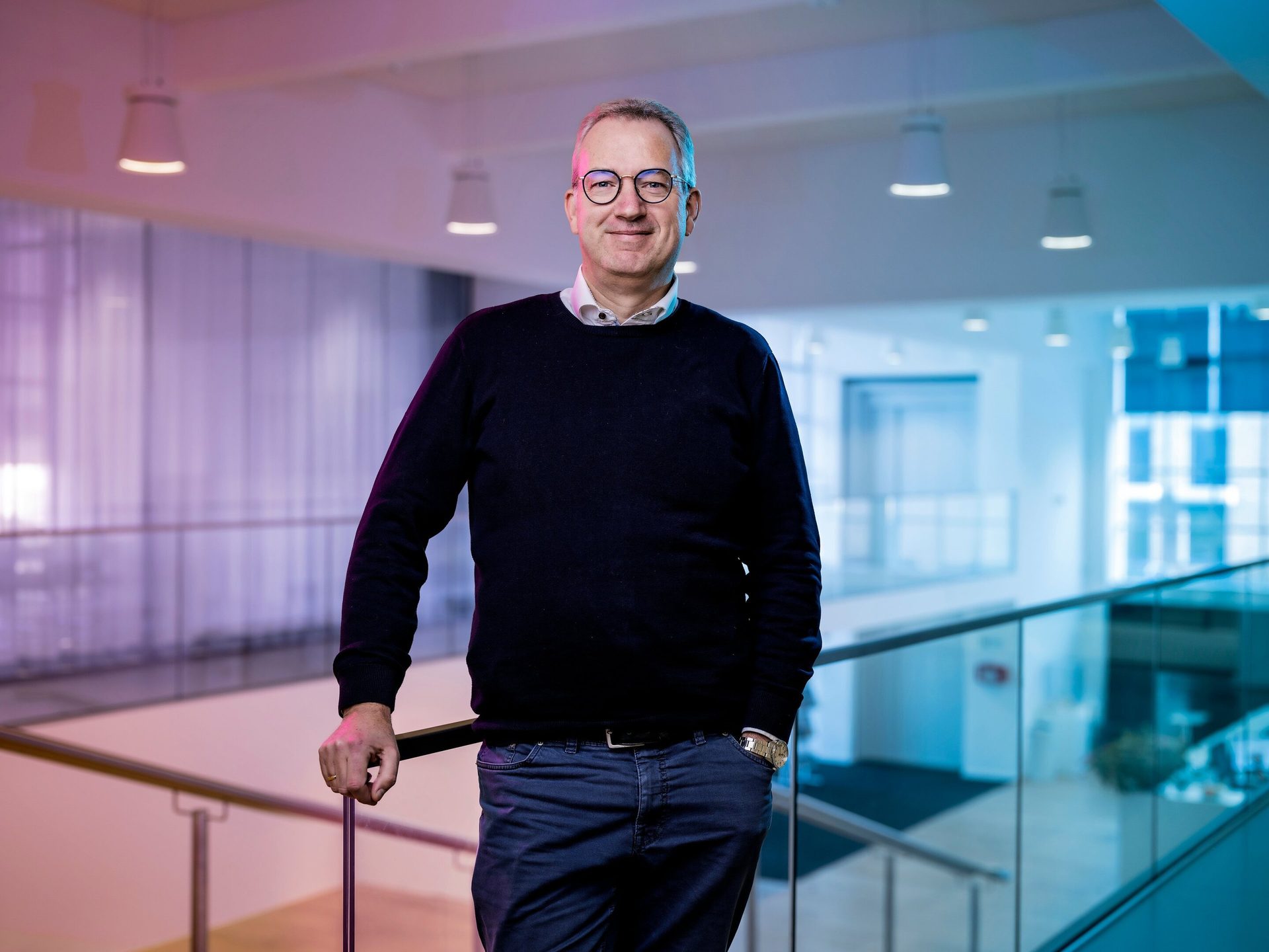 Middle-aged man in glasses, dark sweater, leaning on a railing in a modern, color-lit building.