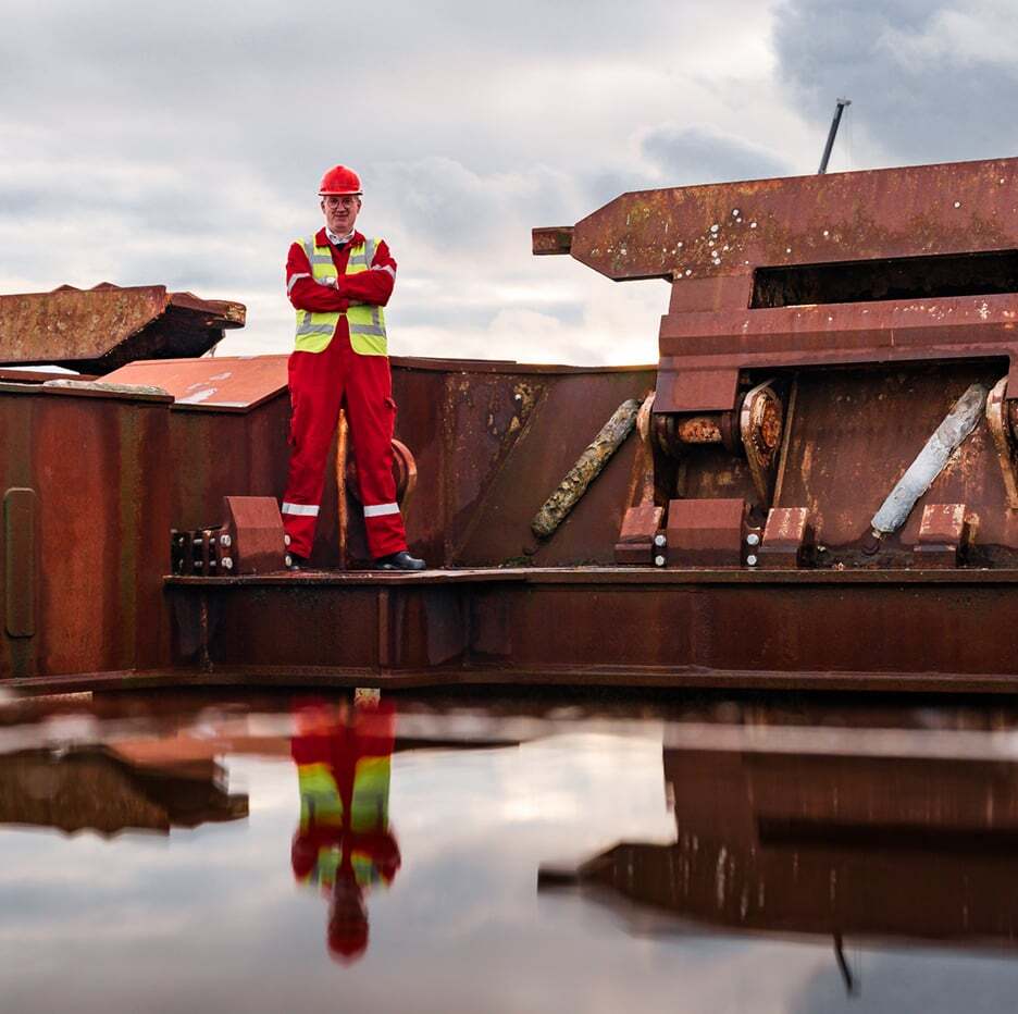 Worker in red safety uniform on rusty structure, reflected in water under cloudy sky.