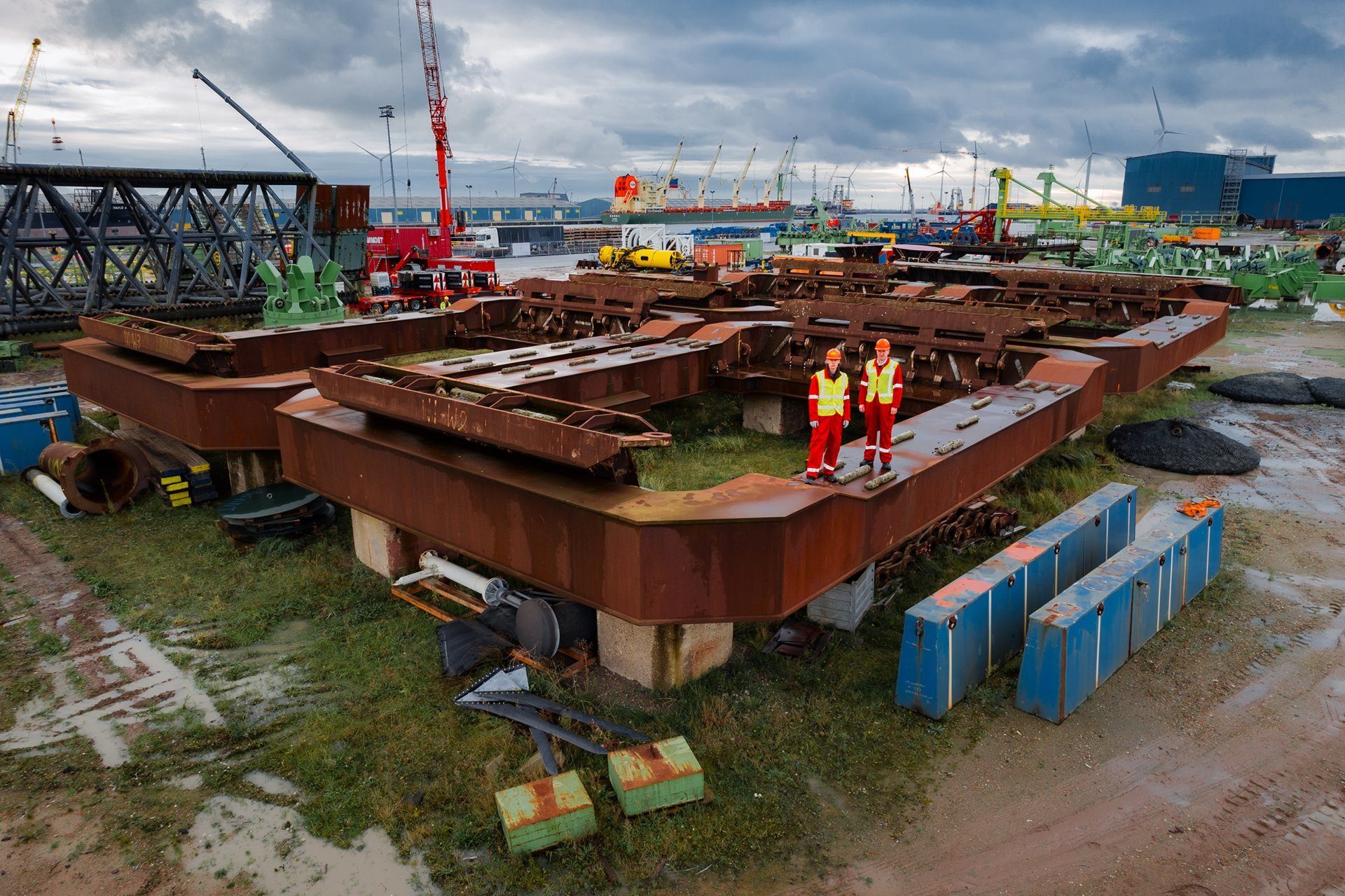 Two workers in safety gear stand on large rusty industrial structures at a muddy port.