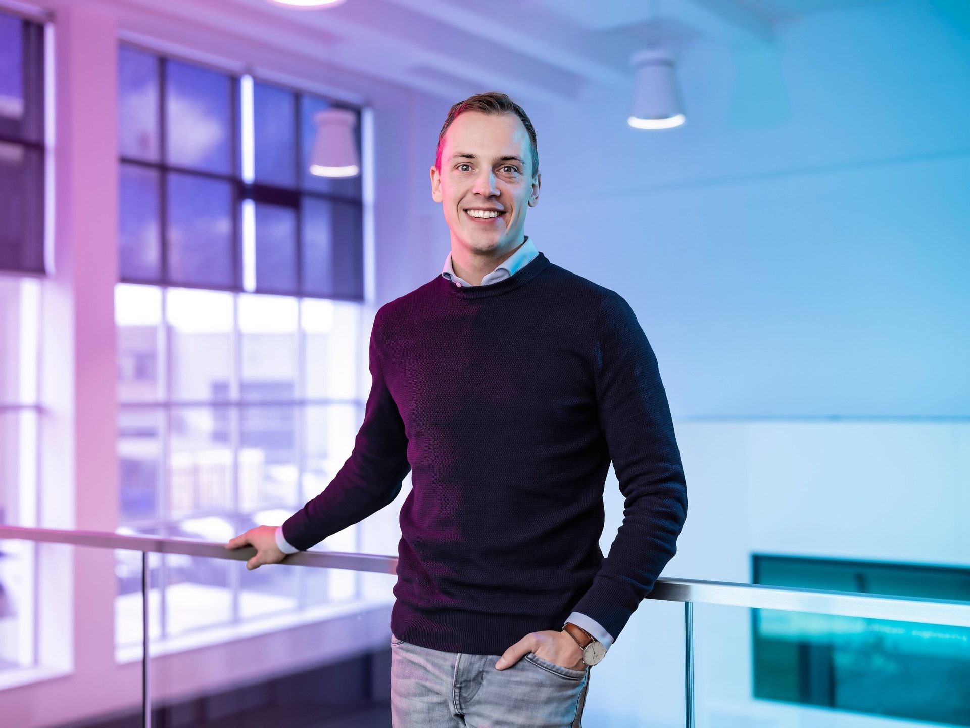 Smiling man leaning on a railing in a modern building with colorful lights.