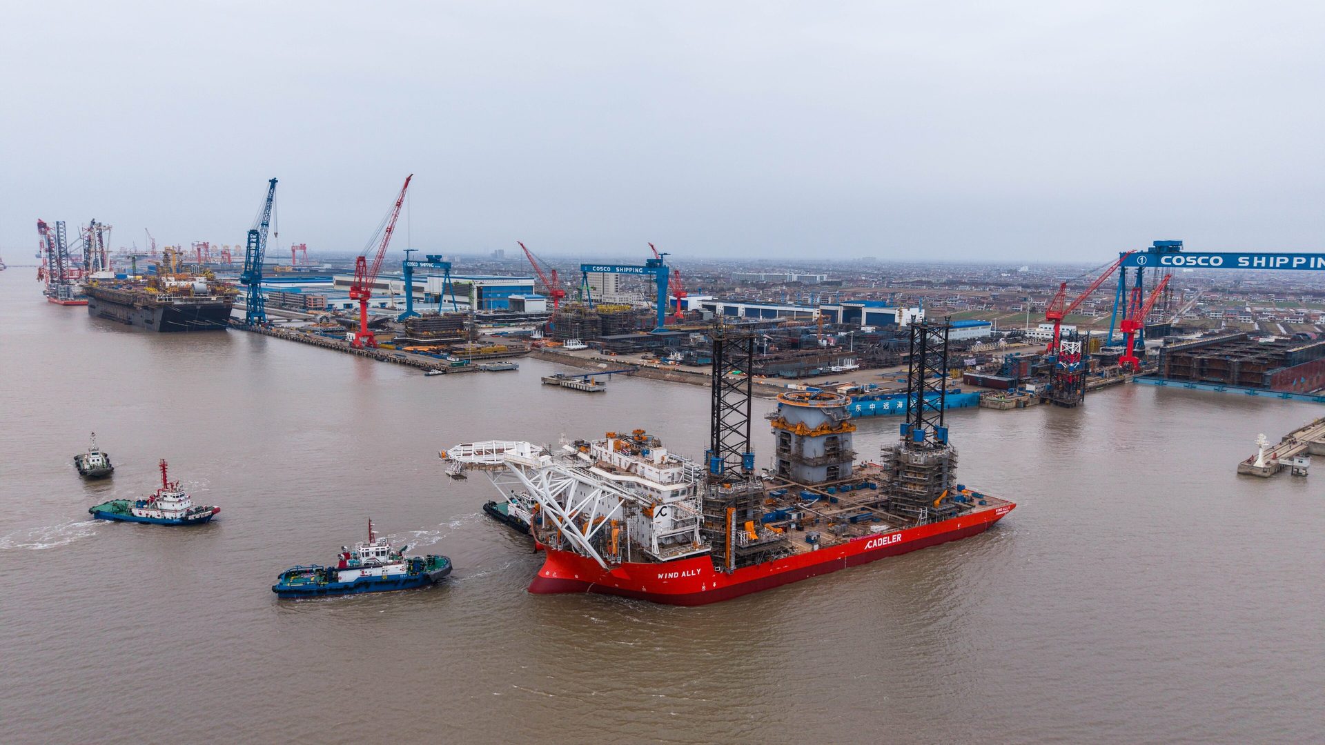 Red offshore wind installation vessel moved by tugboats in a busy shipyard under a gray sky.