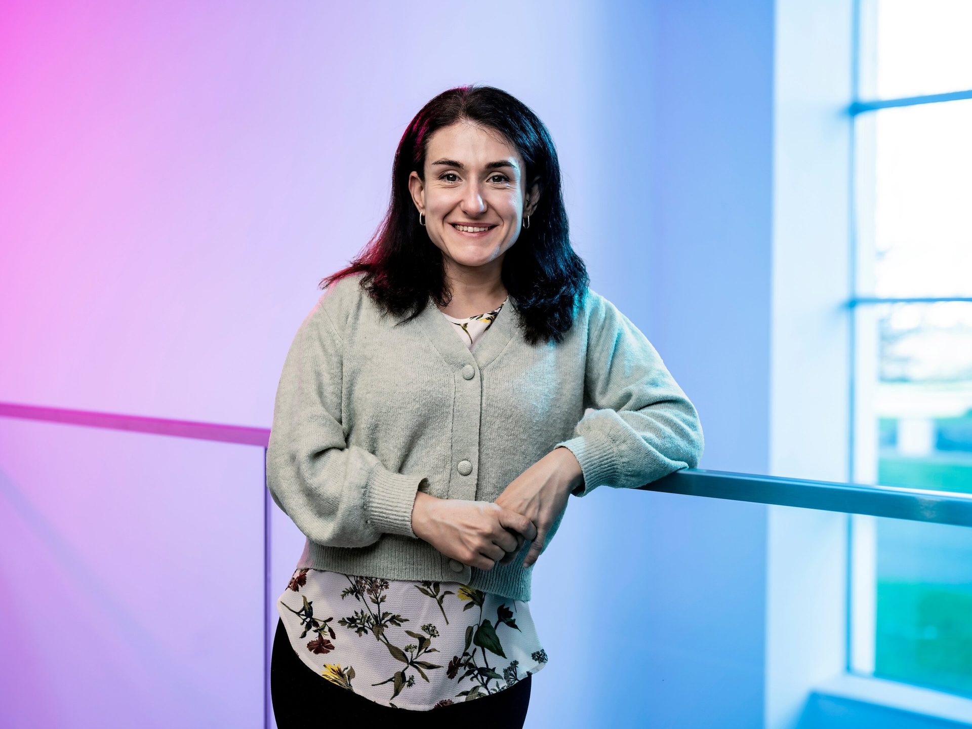Smiling woman in green cardigan leans on a railing against a pink-blue gradient background.