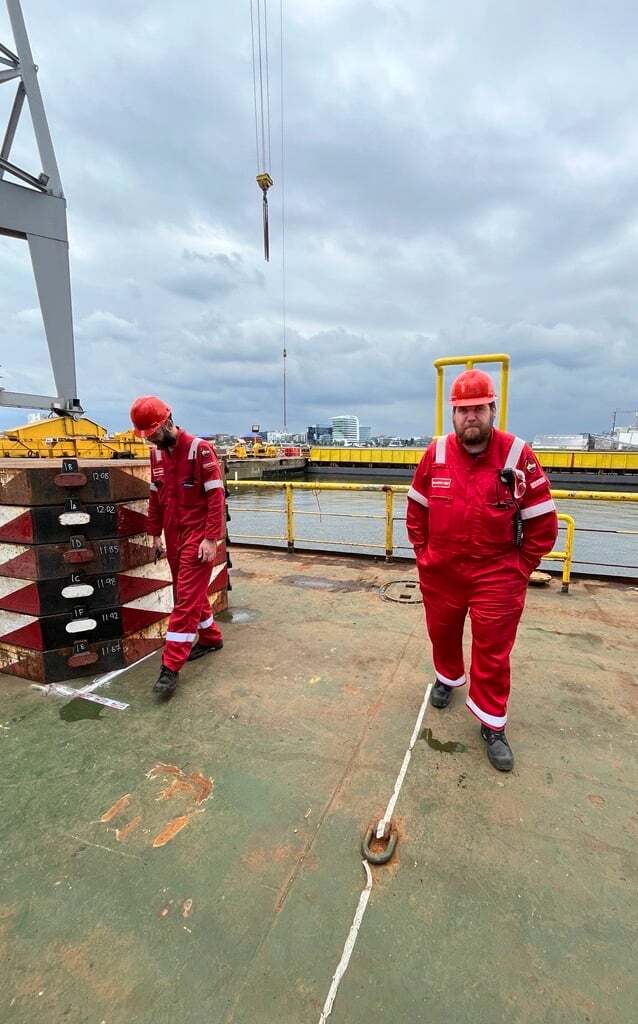 Two workers in red safety gear on a ship deck with a crane overhead and weights.